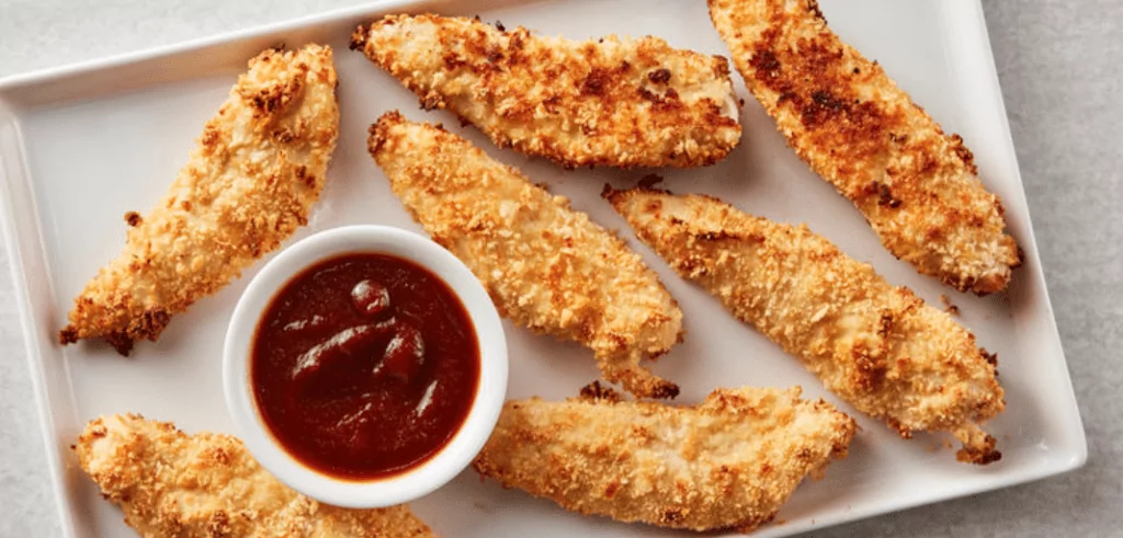 Oven-friend chicken tenders on a white rectangular platter with a red sauce in a ramekin